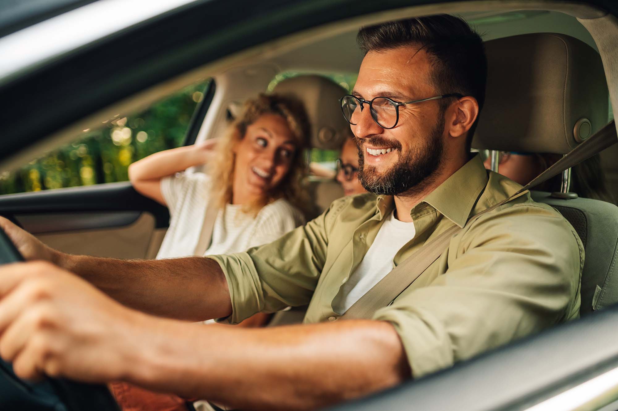 Smiling man wearing glasses driving a car with two passengers in the back seat during daytime.